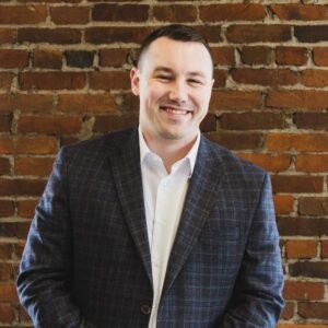 Smiling man in a white shirt and dark plaid blazer posing in front of a brick wall.