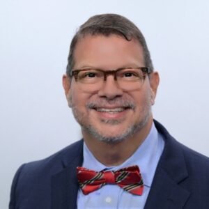 Portrait of a smiling man wearing glasses, a navy suit, light blue shirt, and a red striped bow tie.